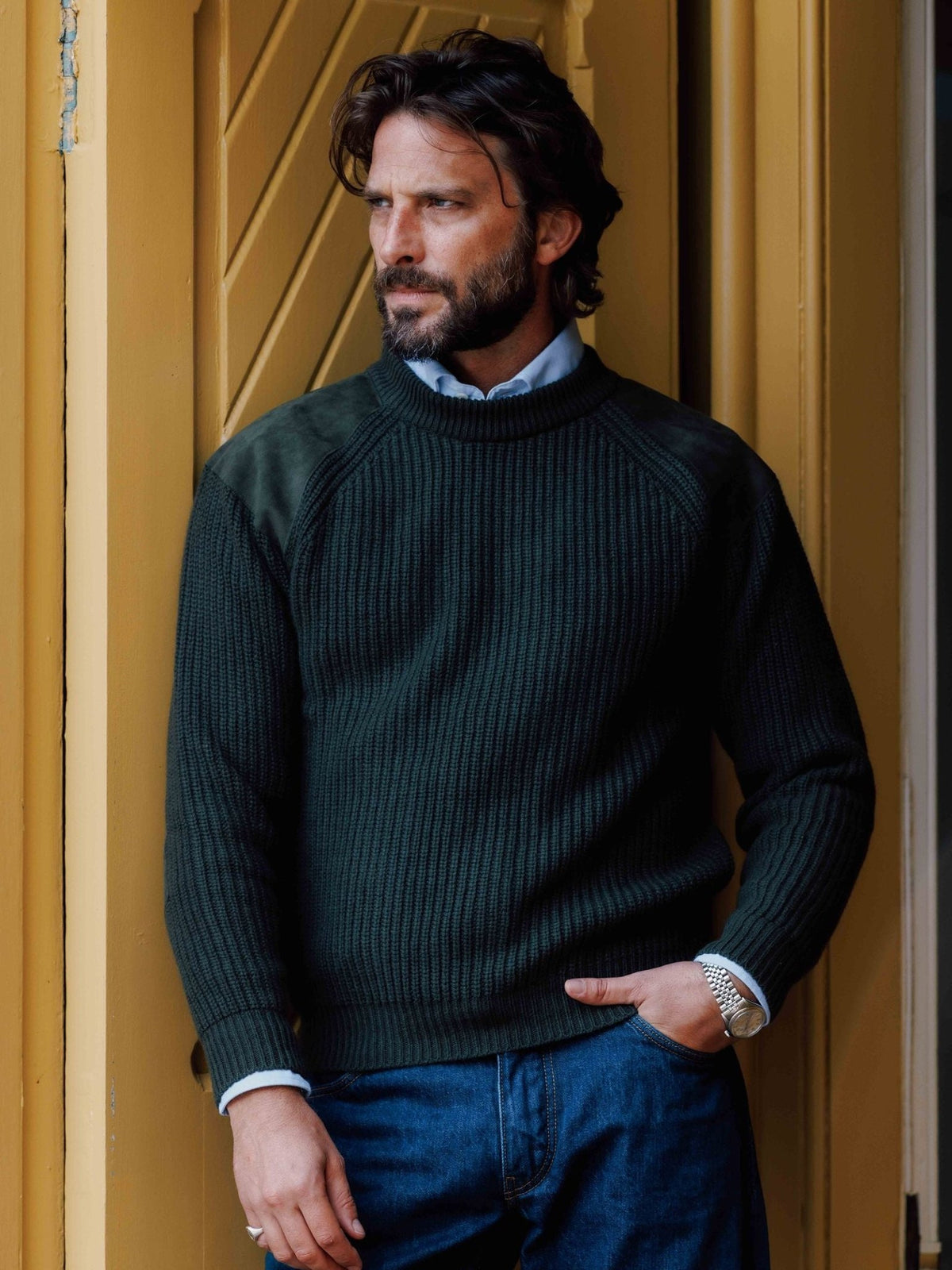 A man wearing a dark green ribbed Campbell’s of Beauly Lux Cashmere Shooter Crew Jumper with tonal shoulder patches over a light blue collared shirt.