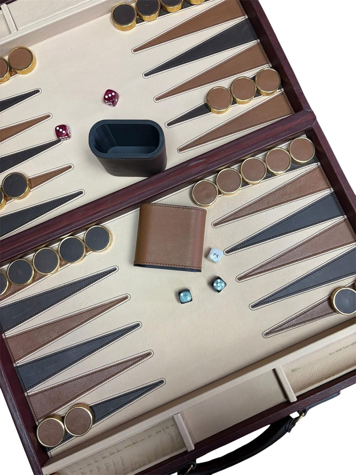 Open the Rey Pavón Leather Backgammon Set to reveal brown, gold, and beige playing pieces arranged on a cream and brown board. Four dice, a matching dice cup, and a brown wallet rest on the board’s surface. A glimpse of the carrying case showcases the refined craftsmanship of this elegant traditional game.