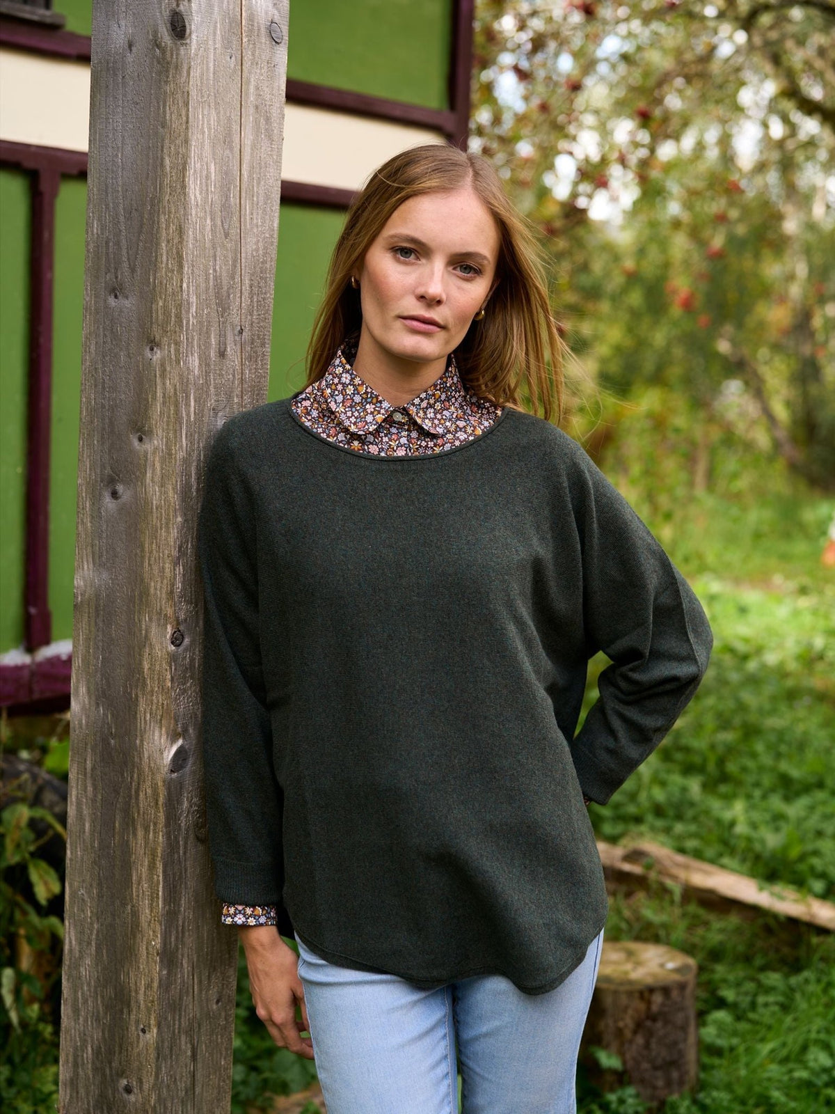 A woman wearing a dark green Merino Cashmere Boat Neck Jumper over a floral print shirt and light blue jeans.