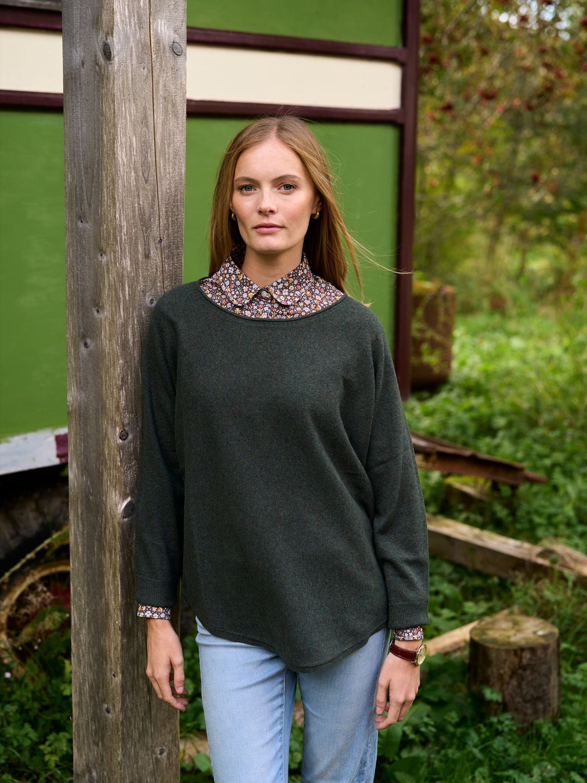 A woman wearing a dark green Campbell’s of Beauly Merino Cashmere Boat Neck Jumper over a floral collared shirt and light blue jeans, standing outdoors next to a wooden post.