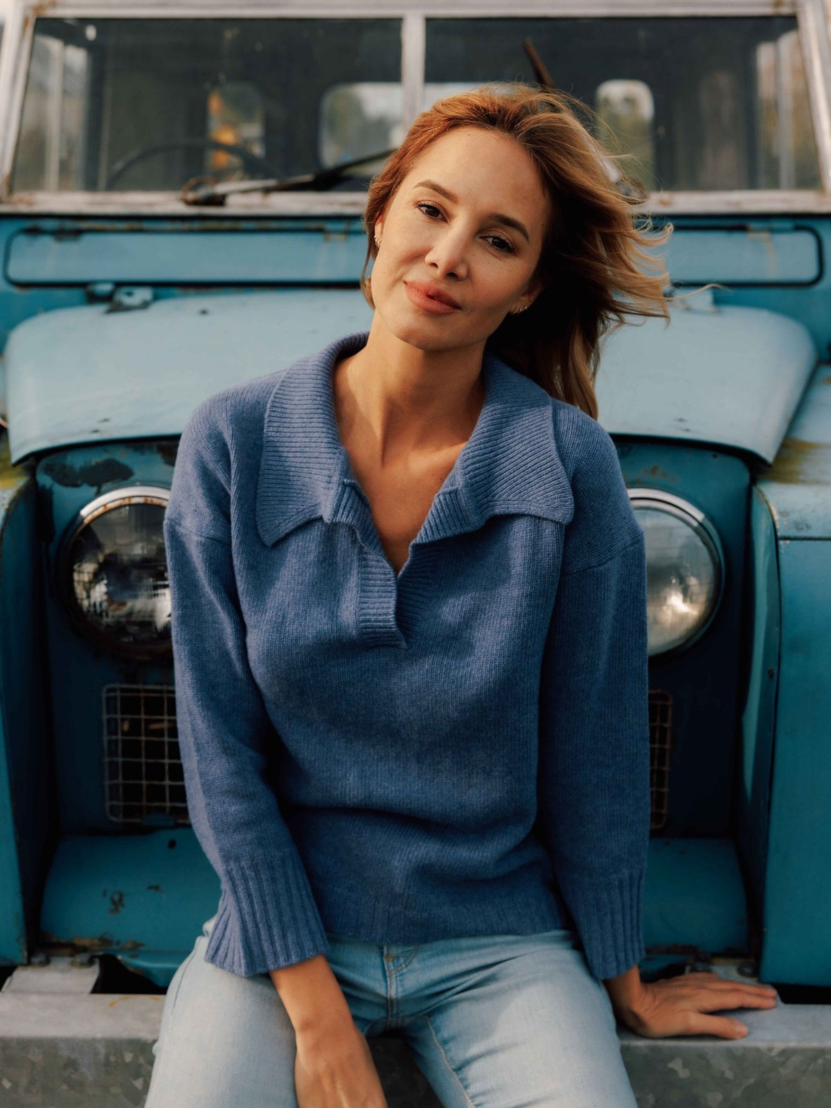 A woman wearing a blue Merino Cashmere Shirt Neck Jumper sits on the front bumper of a vintage blue truck.