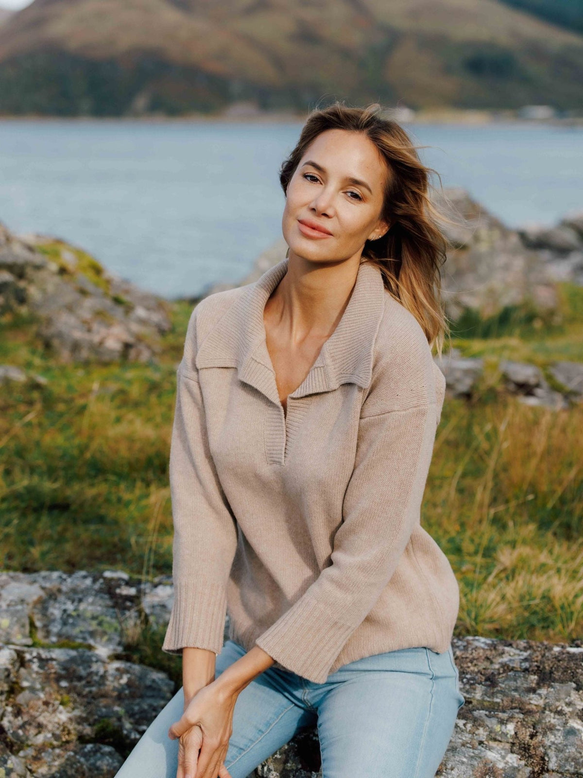 A woman wearing a beige Merino Cashmere Shirt Neck Jumper and light blue jeans, sitting on a rocky shore near a body of water.