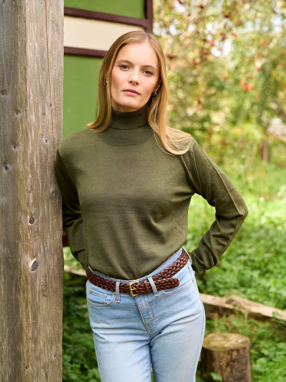 A woman wears a Campbell’s of Beauly Merino Poloneck in olive green paired with light wash jeans and a brown braided belt.