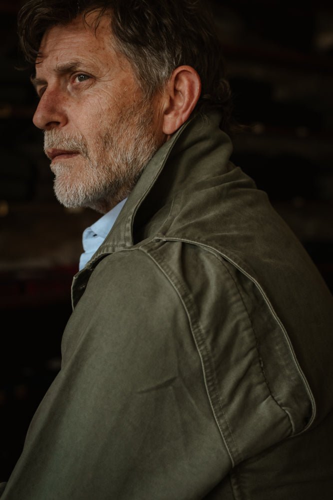 A man wears a Campbell’s of Beauly Nevis Jacket in olive green cotton moleskin with a high collar over a light blue collared shirt.