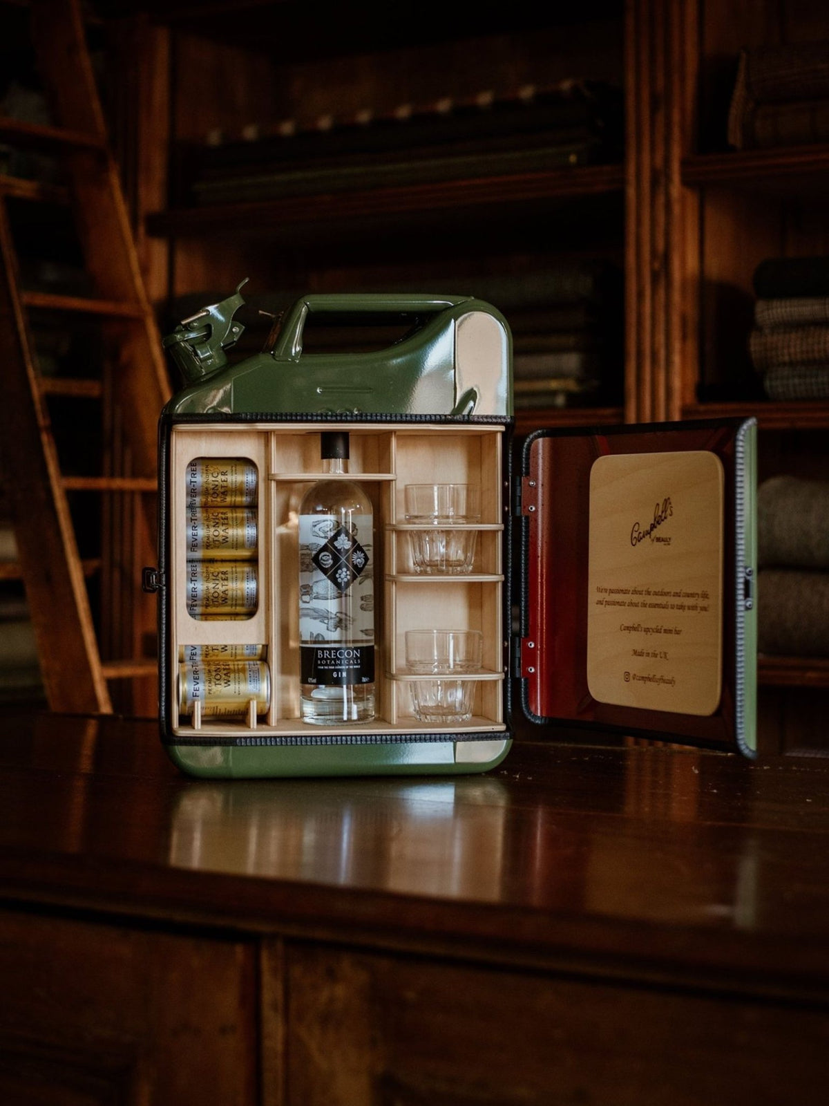 An Original Upcycled Mini Bar made from an open green metal repurposed jerry can, containing a bottle of gin, two glass tumblers, and several cans of tonic water.
