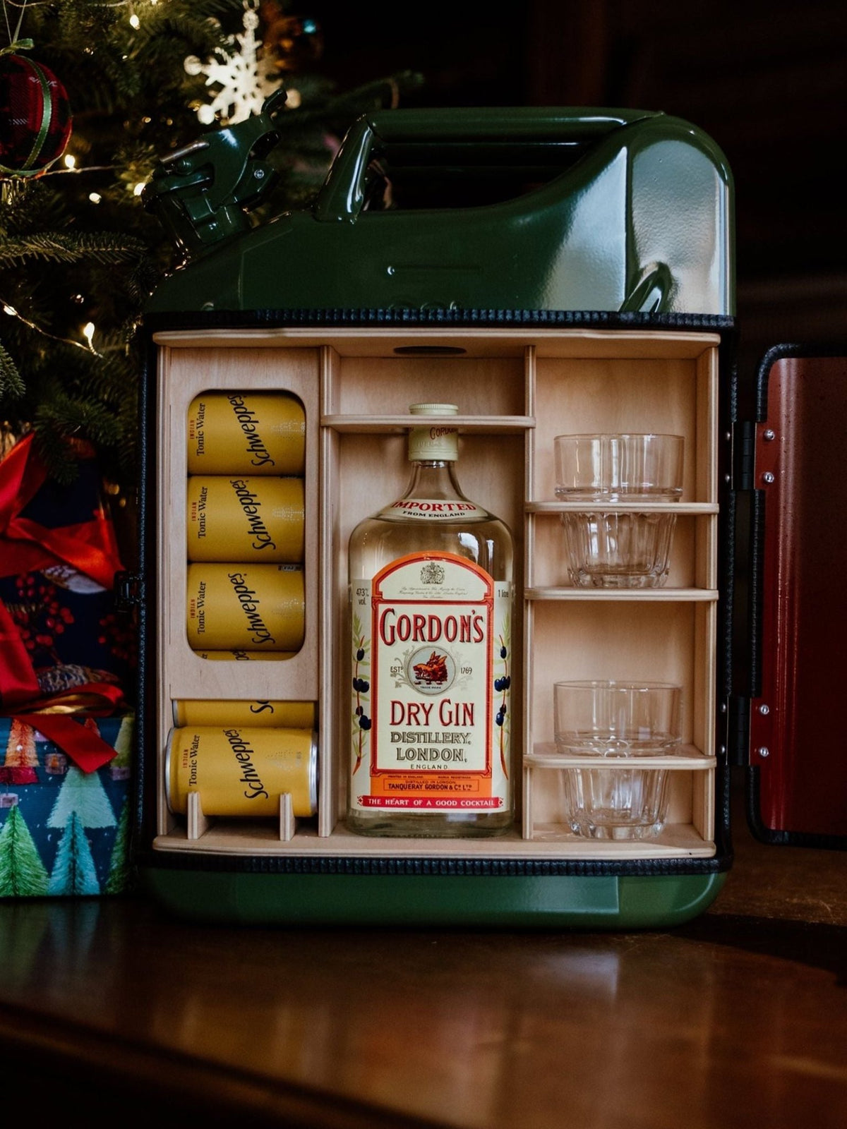 An Original Upcycled Mini Bar made from a green metal jerry can, containing a bottle of gin, four cans of tonic water, and two drinking glasses.