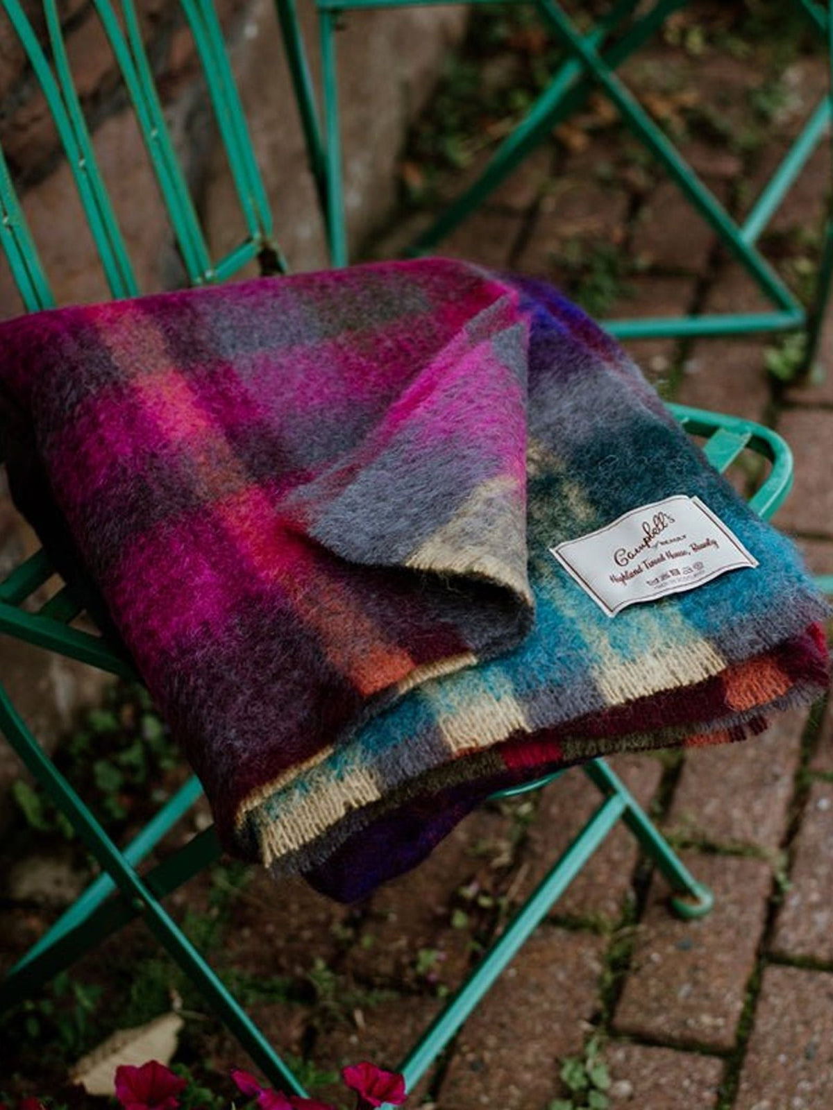 A Painterly Mohair Throw by Campbell’s of Beauly, featuring a colorful plaid pattern and branded label, rests on a green metal chair outdoors beside another empty chair and a brick path dotted with fallen leaves and flowers.