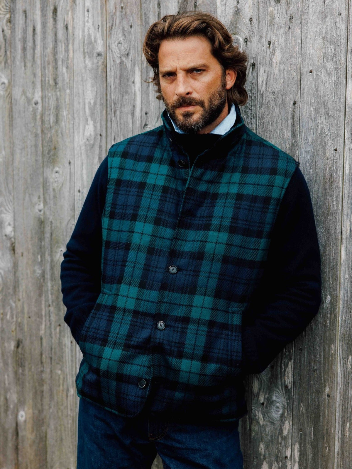 A bearded man leans against a wooden wall wearing a navy tartan Campbell’s of Beauly Quilted Buttoned Gilet over a dark sweater.