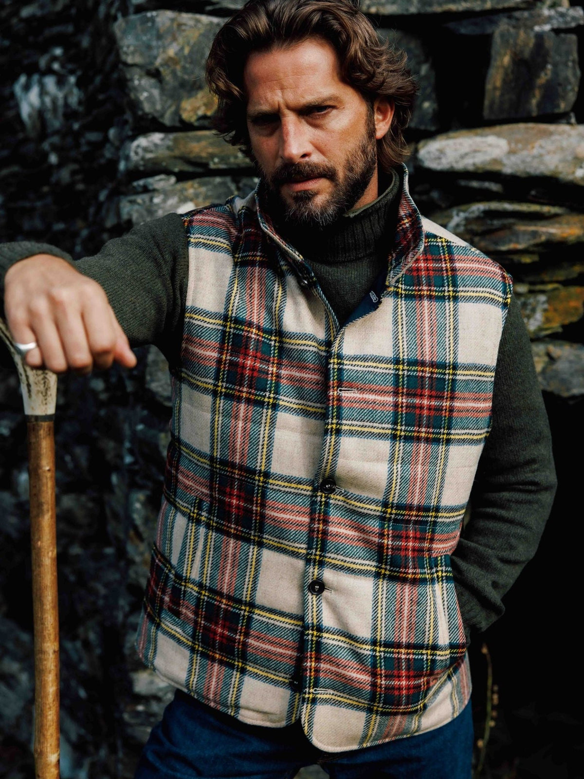 A man wearing a tan and navy tartan Quilted Buttoned Gilet by Campbell’s of Beauly over a dark green turtleneck sweater, standing with a wooden walking stick.