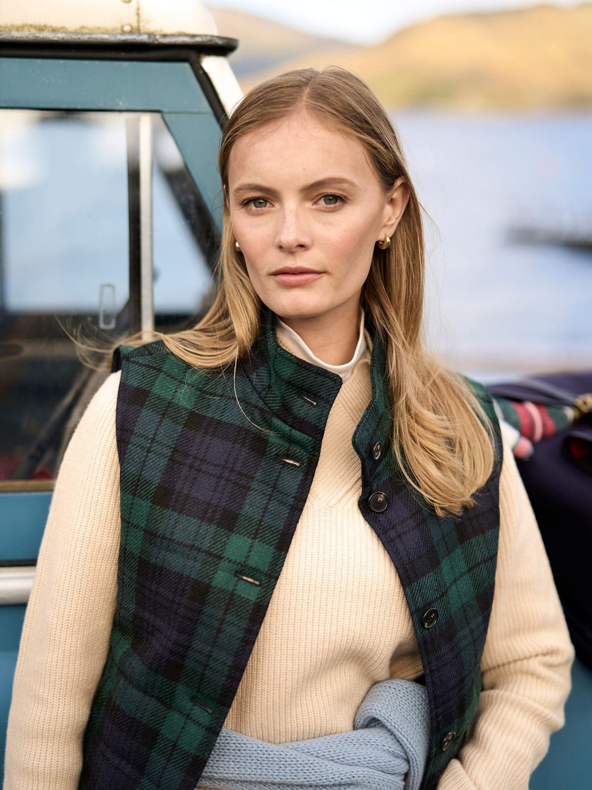 A person wears a Campbell’s of Beauly Quilted Buttoned Tartan Gilet in Antique Black Watch over a cream-colored sweater.