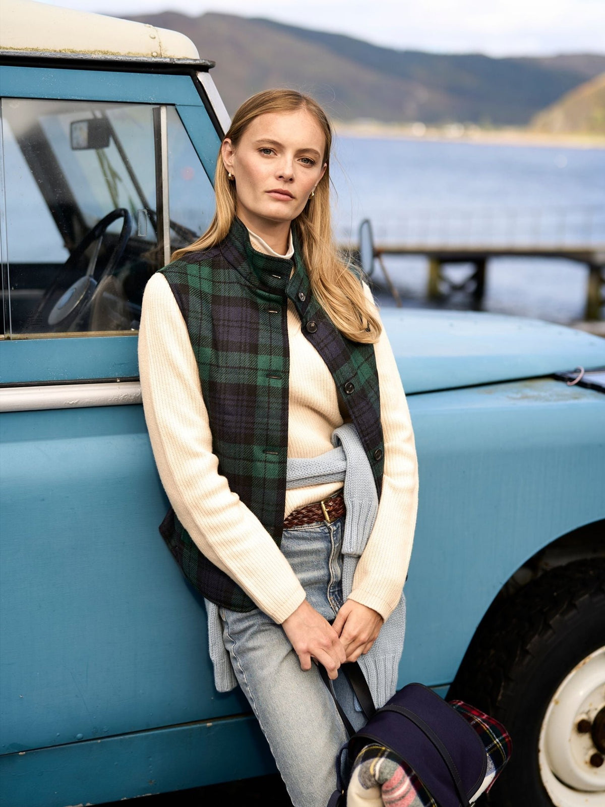 A woman wearing a Campbell’s of Beauly Quilted Buttoned Tartan Gilet in Antique Black Watch over a cream sweater, leaning against a blue vintage vehicle.