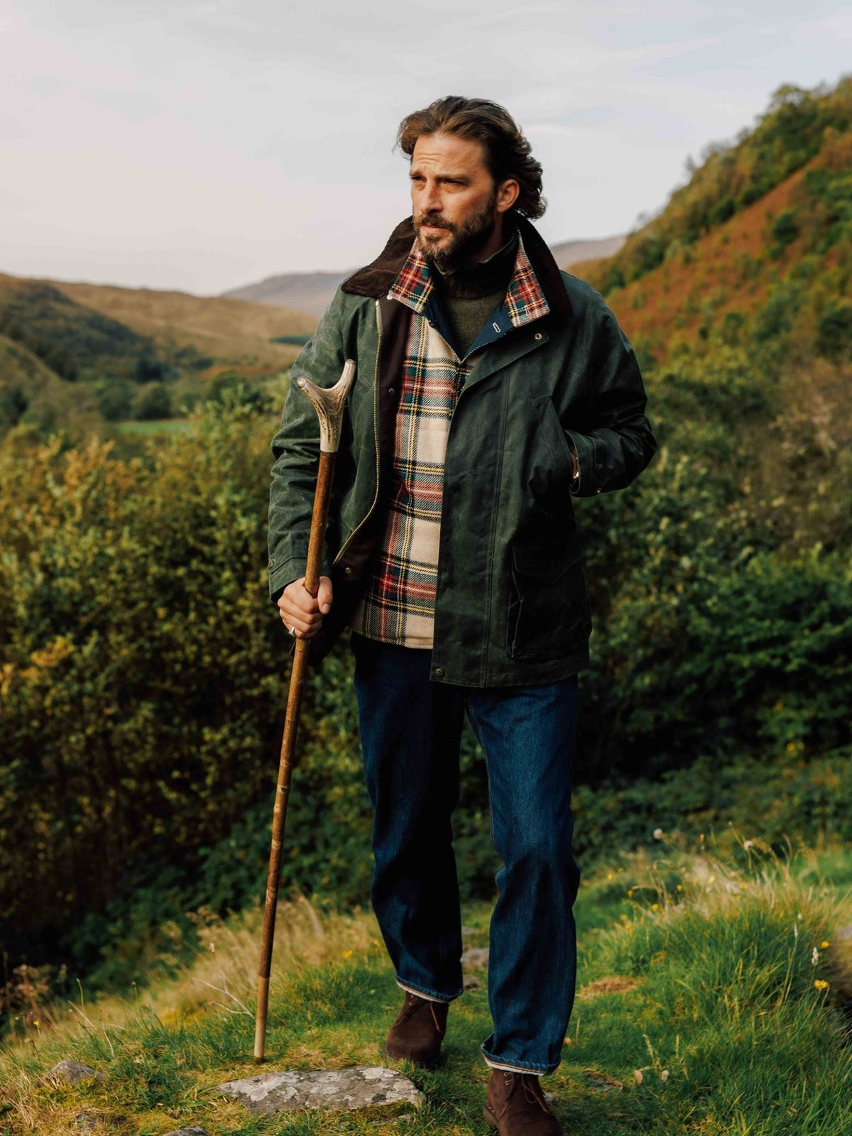 A man wearing a green waxed cotton Rain Cabaan by Campbell’s of Beauly with a plaid lining and dark jeans stands on a hillside holding a wooden walking stick.