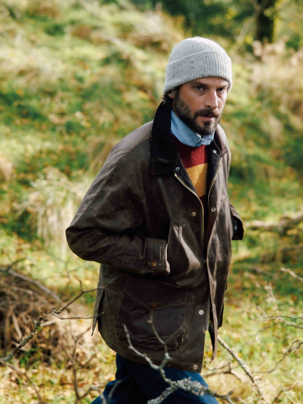 A man wears a grey beanie, a multi-colored sweater under a blue shirt, and a brown Campbell’s of Beauly Rain Cabaan heritage waxed cotton jacket outdoors.