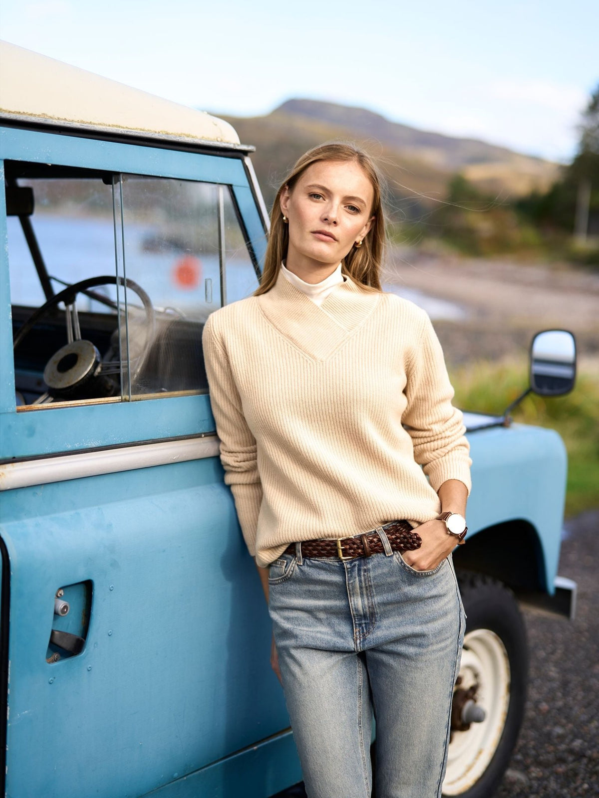 A woman wears a cream ribbed merino wool Rib V Neck Jumper by Campbell’s of Beauly over a white turtleneck while leaning against a blue vintage vehicle.