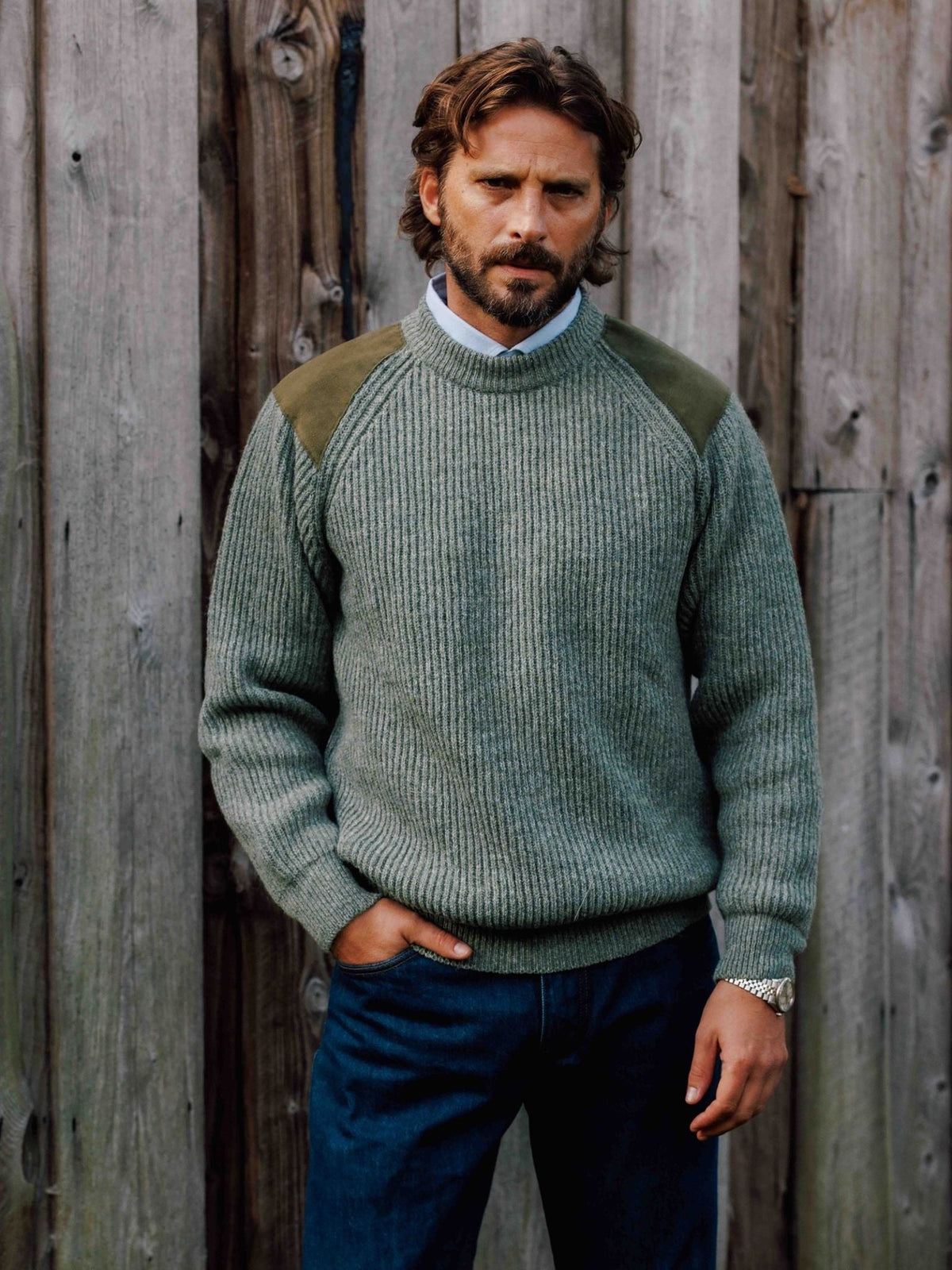 A man wears a green ribbed knit Scotia Suede Jumper with contrasting shoulder patches over a light blue collared shirt.