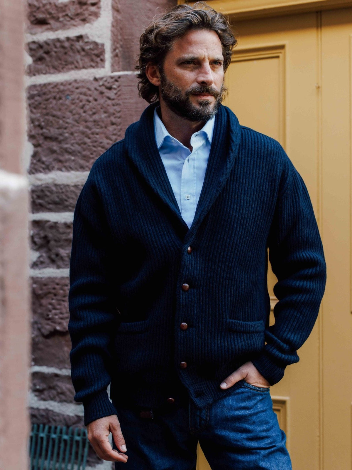 A man wearing a navy blue Campbell’s of Beauly Shawl Collar Cardigan over a light blue button-down shirt.