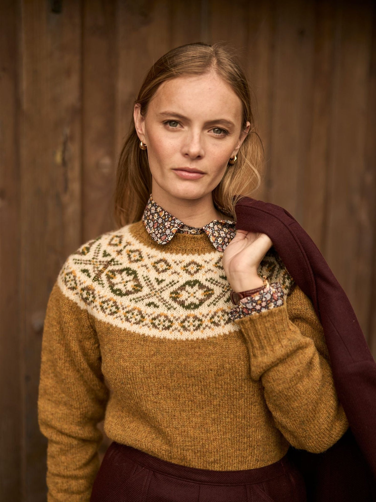 A woman wears a mustard-colored Campbell’s of Beauly Shetland Fairisle Crew with a floral print collar over a dark skirt, with a dark jacket draped over her shoulder.
