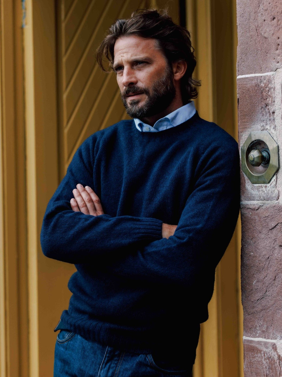 A bearded man wears a blue button-down shirt under a navy blue Campbell’s of Beauly Shetland Jumper as he leans against a stone wall with his arms crossed.