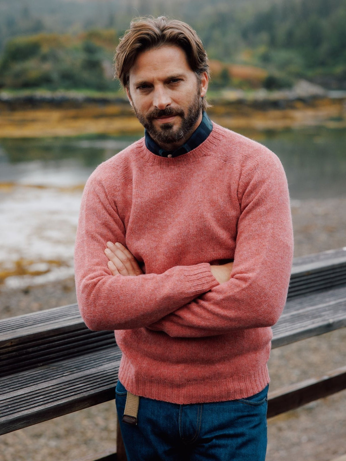 A man with brown hair and a beard stands outdoors by a wooden railing, wearing the Campbell’s of Beauly Shetland Jumper in pink with blue jeans, his arms crossed. Trees and water are blurred in the background.