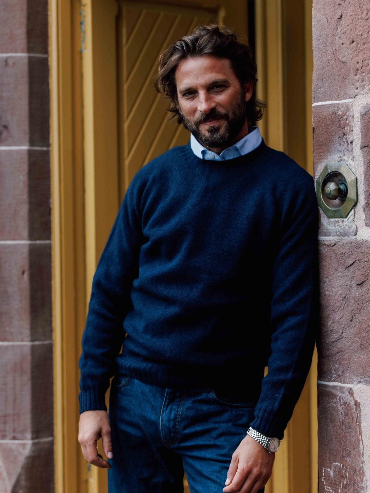 A man wearing a navy blue Campbell’s of Beauly Shetland jumper over a light blue collared shirt and blue jeans, leaning against a stone wall.