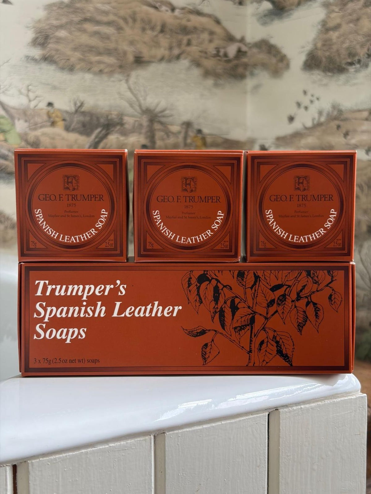 Three orange boxes of Geo. F. Trumper Spanish Leather Hand Soap, Set of 3, are arranged on a matching illustrated box against a white surface and nature-themed wallpaper backdrop.