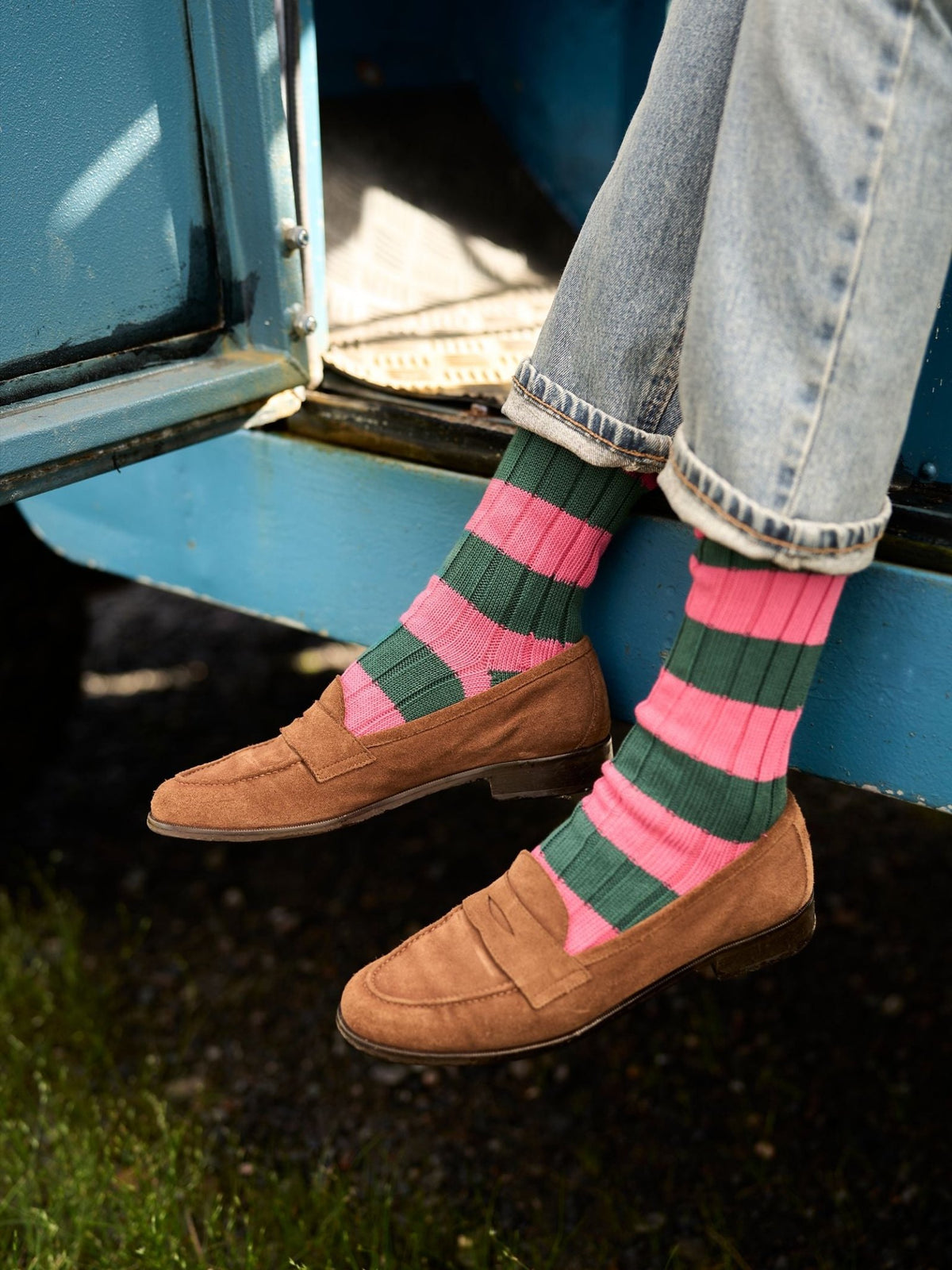 Person wearing pink and green Stripe Socks and brown suede loafers, sitting on the edge of a blue vehicle.