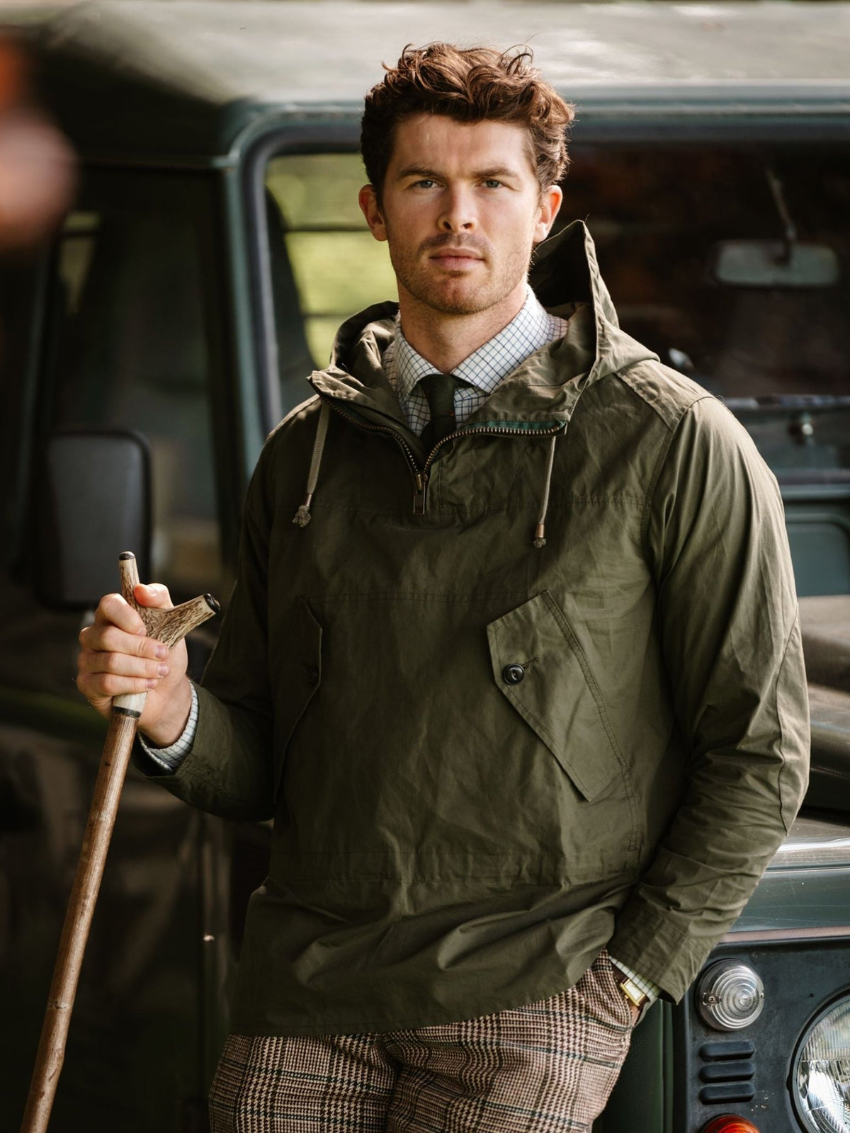 A man wearing a green Campbell’s of Beauly The Stalker Smock over a dress shirt and tie, leaning against a vehicle and holding a wooden walking stick.