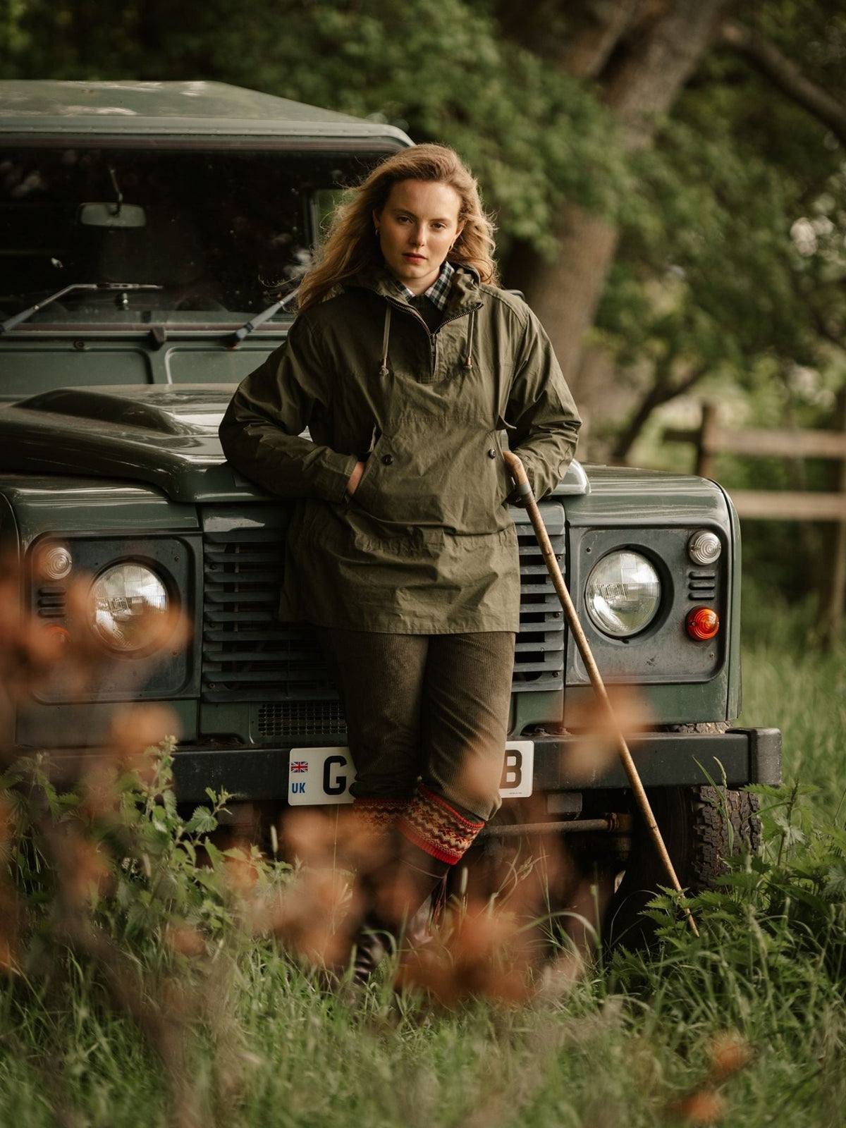 A person wearing an olive green dry wax ripstop Stalker Smock by Campbell’s of Beauly and wool breeks leans against the front of a dark green off-road vehicle, holding a wooden walking stick.