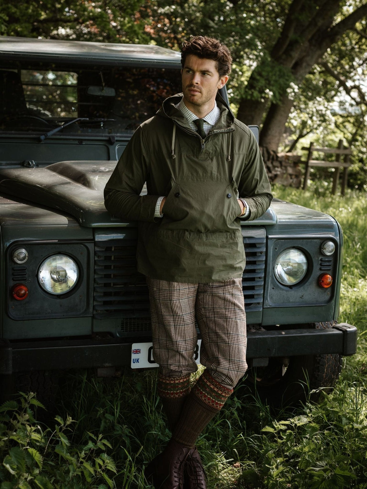 A man wearing an olive green Campbell’s of Beauly The Stalker Smock, checkered trousers, and tall patterned socks, leaning against a dark green off-road vehicle.