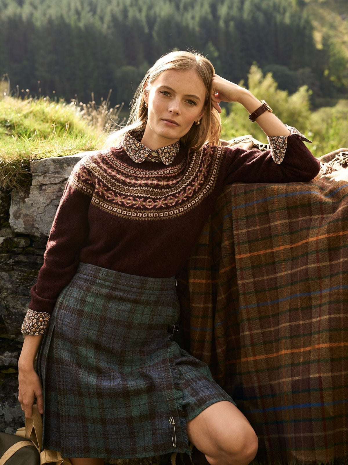 A woman wearing a patterned sweater over a collared shirt and a Campbell’s of Beauly Tweed Kilt, leaning against a stone wall draped with a tartan blanket.