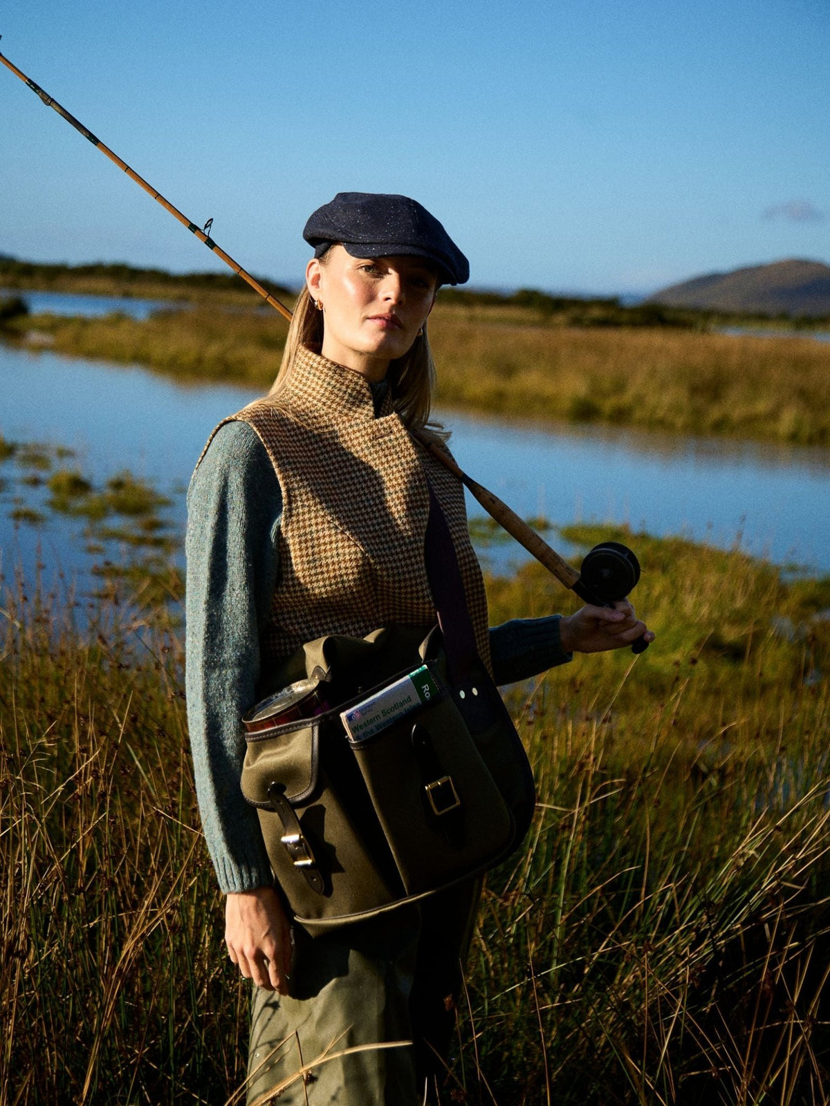 A woman stands in tall grass by a river, wearing the Campbell’s of Beauly Tweed Nehru vest with a cap and sweater, holding a fishing rod and enjoying the outdoors under a clear blue sky.