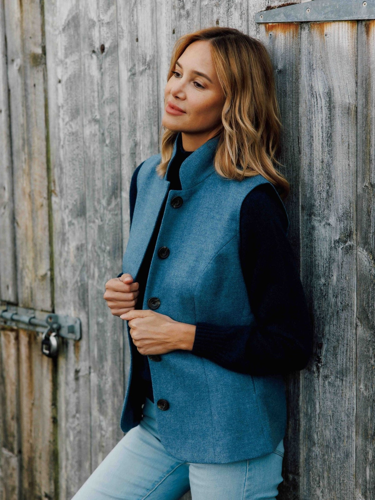 A woman wearing a Campbell’s of Beauly Tweed Nehru gilet over a black sweater, leaning against a weathered wooden gate.