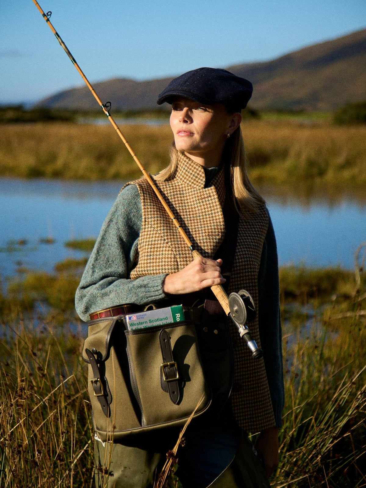 A woman stands by a lake, wearing the Campbell’s of Beauly Tweed Nehru, a cap, green sweater, and country-style shoulder bag marked Welcome to Scotland. Grassy hills and water are behind her.