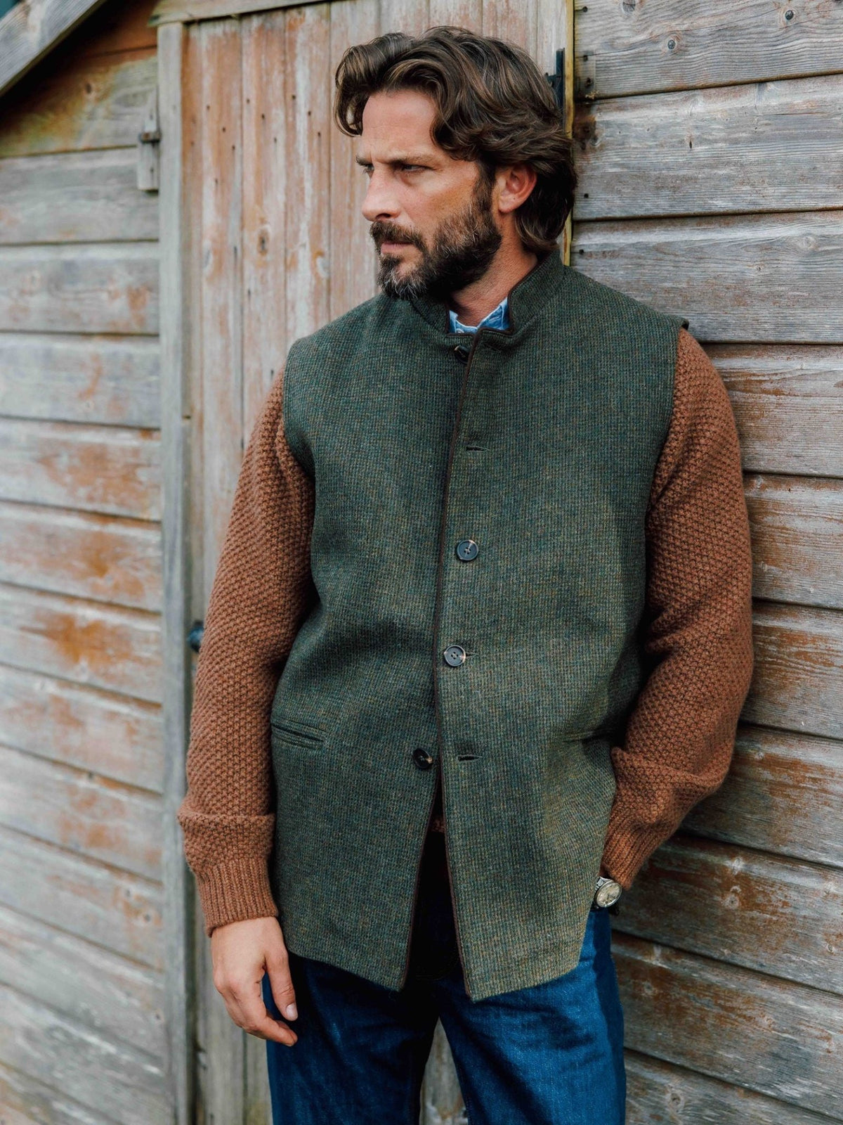 A man wearing a green British Tweed Nehru Gilet by Campbell’s of Beauly over a brown textured sweater, leaning against a wooden wall.