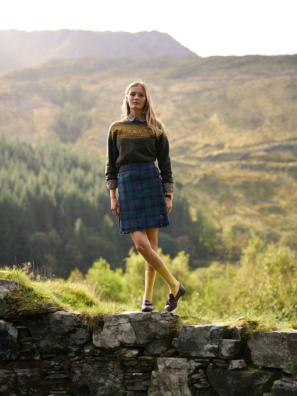 A woman stands on a stone wall wearing a Campbell’s of Beauly Two-Colour Fairisle Yoke Jumper over a collared shirt, paired with a blue and green plaid kilt, yellow socks, and loafers.