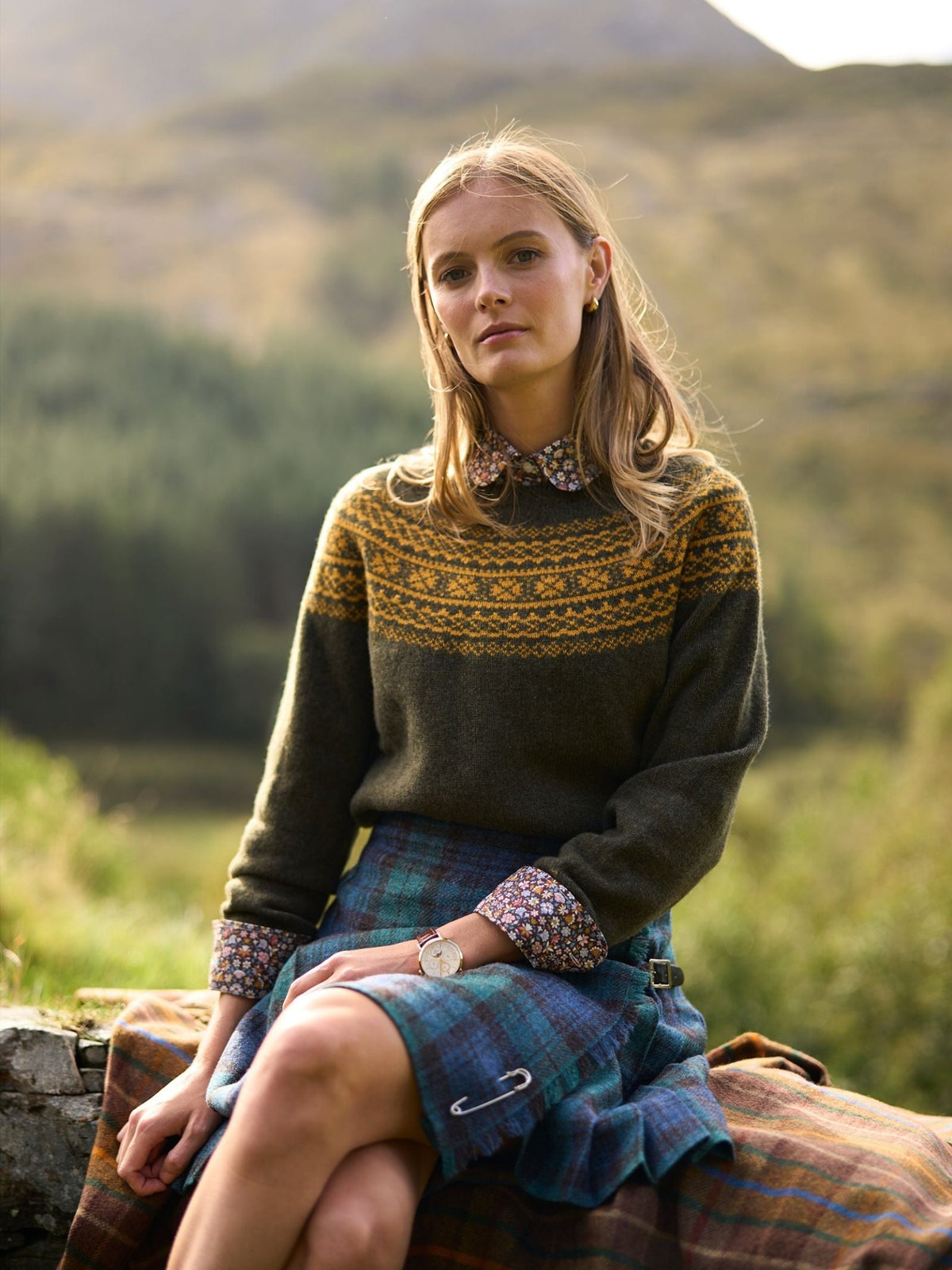 A woman wearing a green Two-Colour Fairisle Yoke Jumper by Campbell’s of Beauly over a floral shirt and a plaid kilt, sitting outdoors in a hilly landscape.