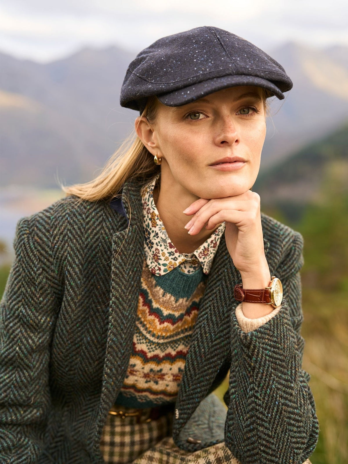A person wearing a dark British Unisex Affric Cap, a patterned sweater, and a herringbone blazer, resting their chin on their hand.