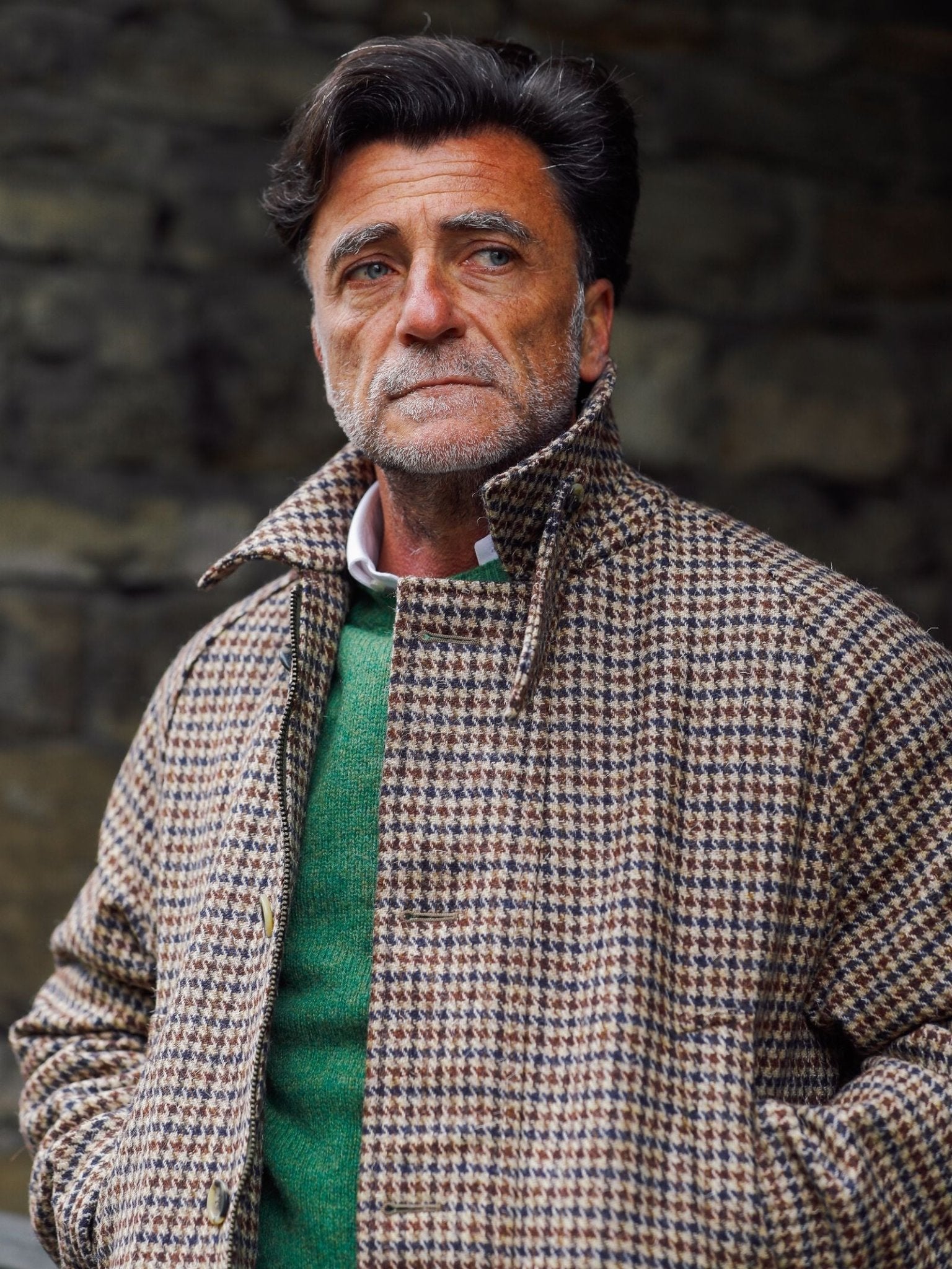A middle-aged man with dark hair and a beard wears the Campbell's of Beauly Cannich Coat over a green sweater, standing outdoors by a stone wall, gazing thoughtfully into the distance.