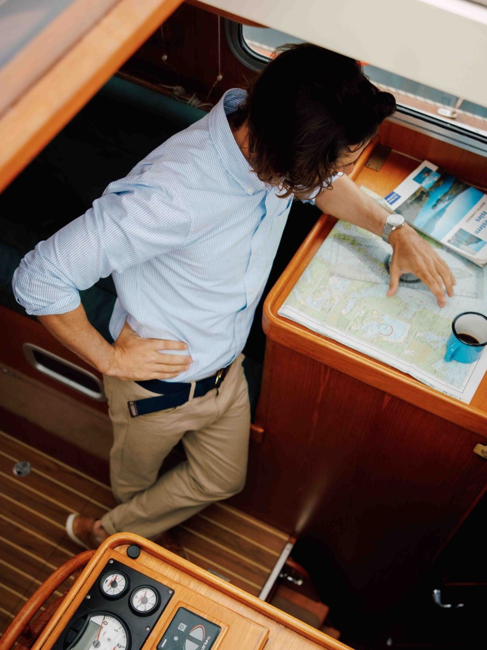 A man in a blue shirt and khaki pants, wearing a Campbell's of Beauly Canvas D-Loop Belt, studies a map on a wooden desk inside a boat, with coffee and navigation instruments nearby.
