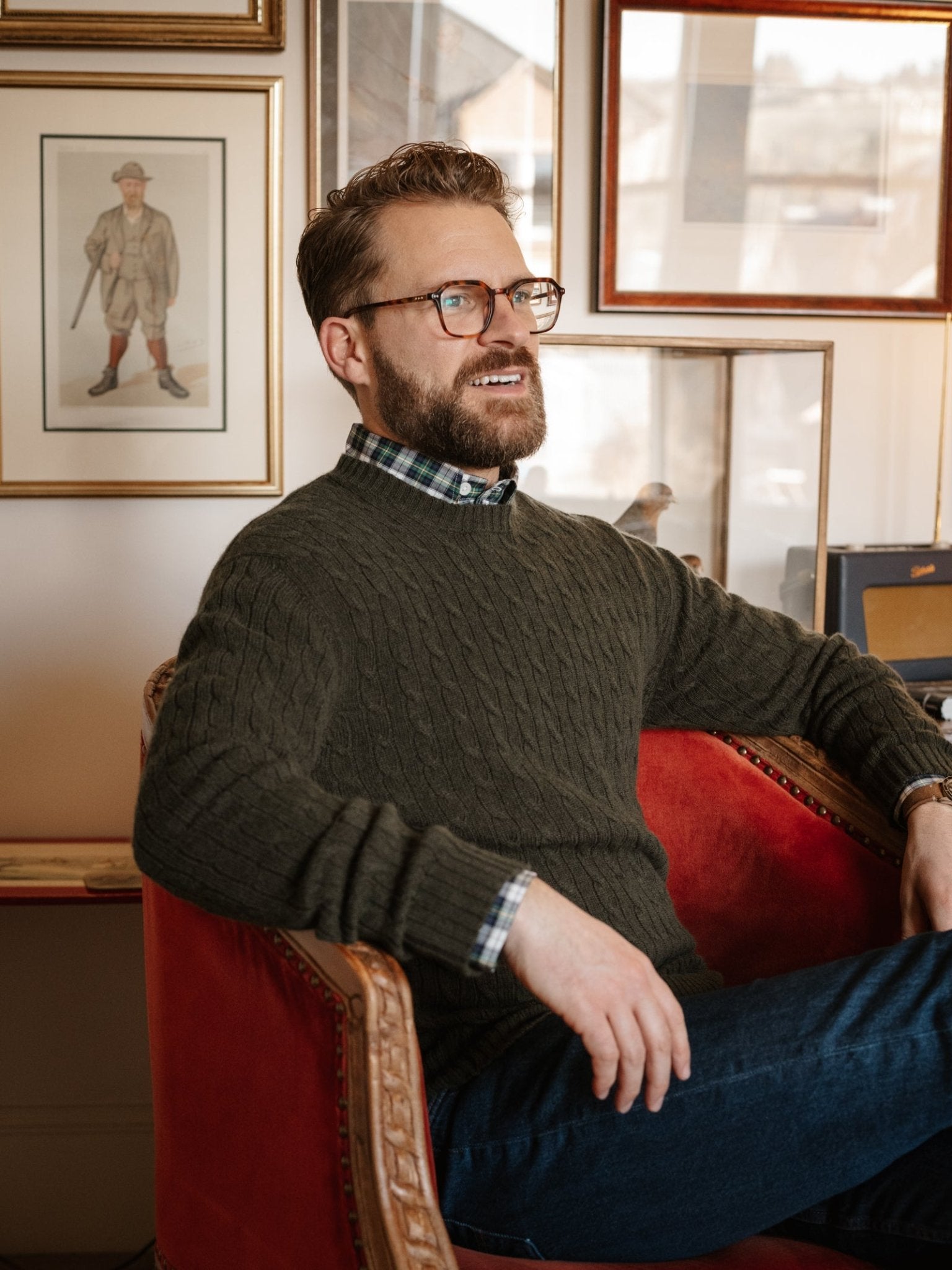A bearded man with glasses, dressed in Campbell's of Beauly Cashmere Cable Crew Jumper and jeans, relaxes on a red upholstered chair in a room decorated with framed art and vintage décor.