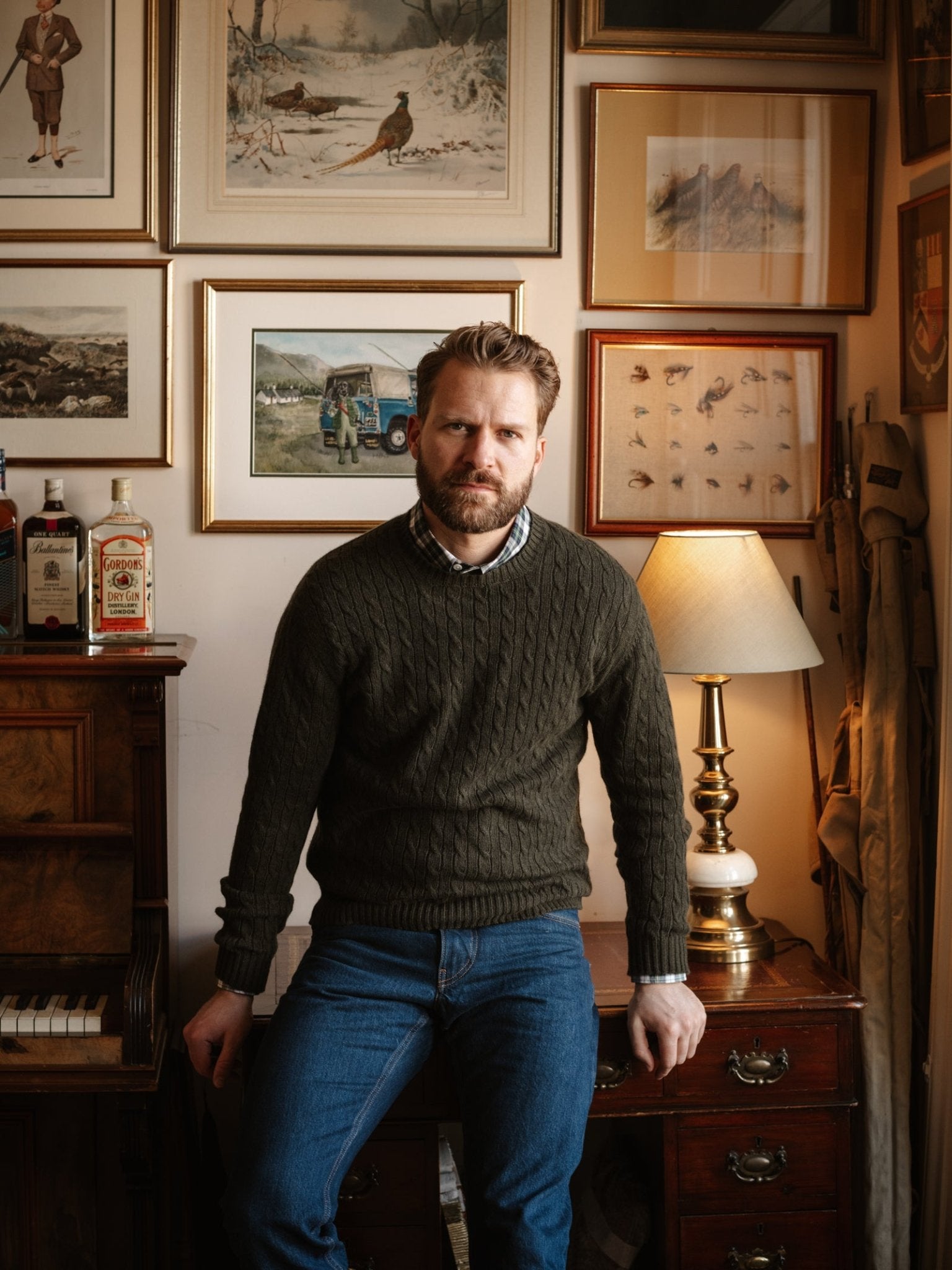 A bearded man in the Campbell's of Beauly Cashmere Cable Crew Jumper and jeans sits on a desk in a warm, vintage room with framed art, bottles, a lamp, and a piano.