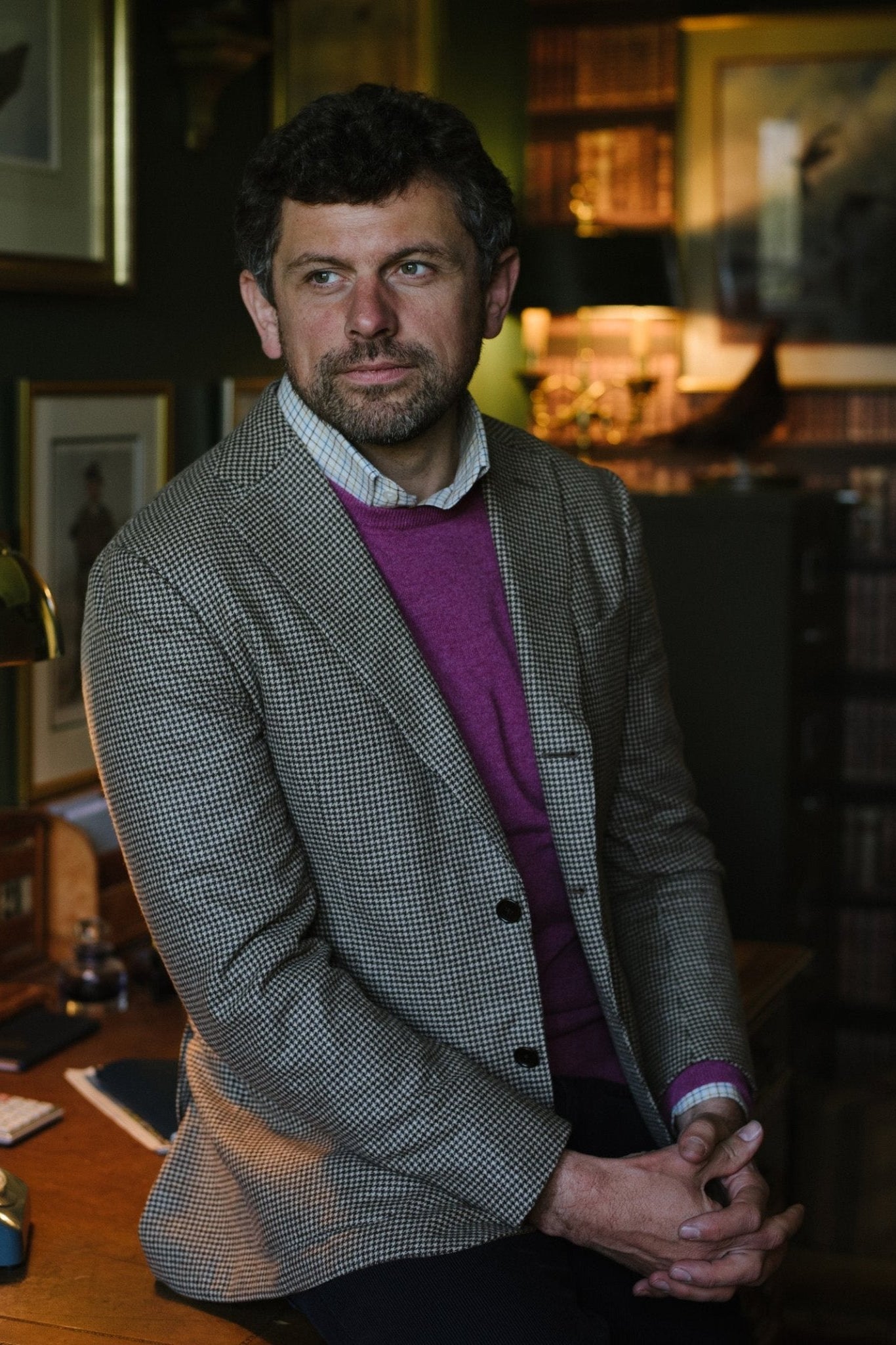 A man with curly hair and a beard, in a checked blazer and a Cashmere Crew Neck Jumper by Campbells of Beauly, sits on the edge of a wooden desk in a warmly lit study lined with books and framed pictures.