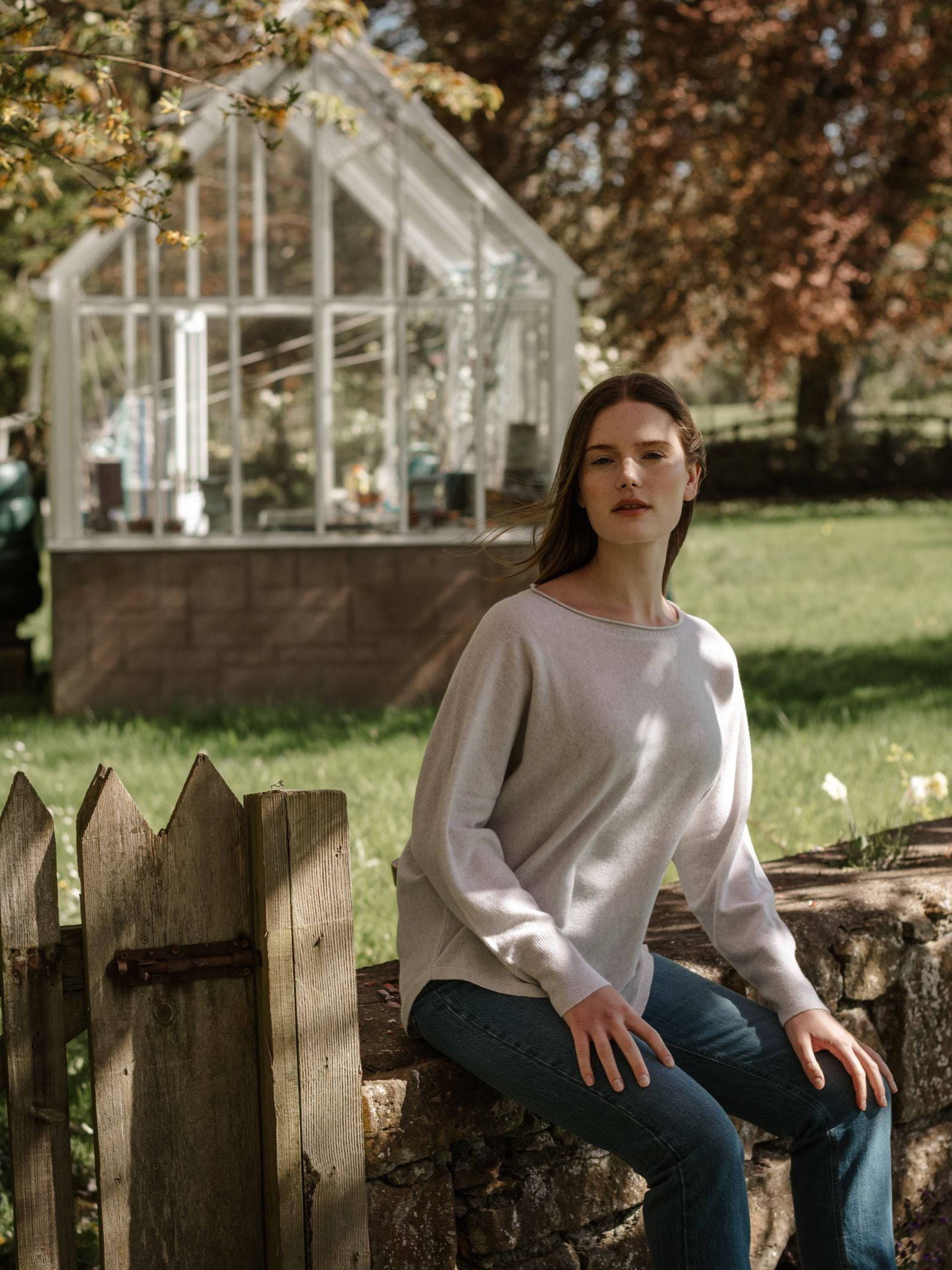 A woman in a Campbell's of Beauly Merino Cashmere Boat Neck sweater and jeans sits on a stone wall by a wooden fence, with a glass greenhouse and green lawn behind her on a sunny day.