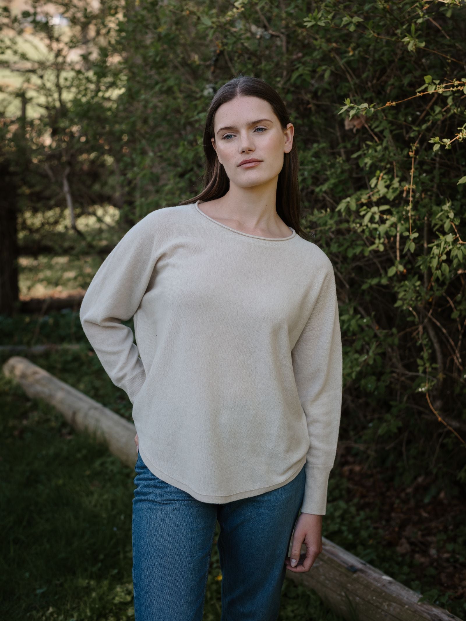 A woman with long brown hair wears the Campbell's of Beauly Merino Cashmere Boat Neck sweater in light beige and blue jeans, standing outdoors by green bushes and trees, calmly looking at the camera with one hand by her side.
