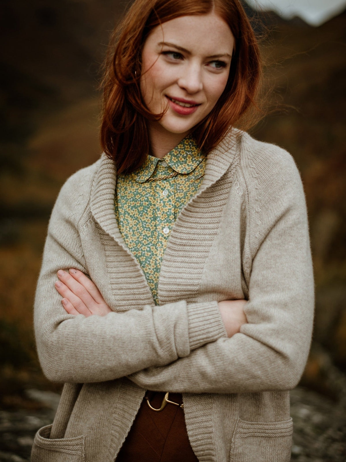 A young woman with long red hair stands outdoors, smiling slightly, wearing the Campbells of Beauly Cashmere Shawl Cardigan over a green floral blouse. Her arms are crossed and the natural background is softly blurred.