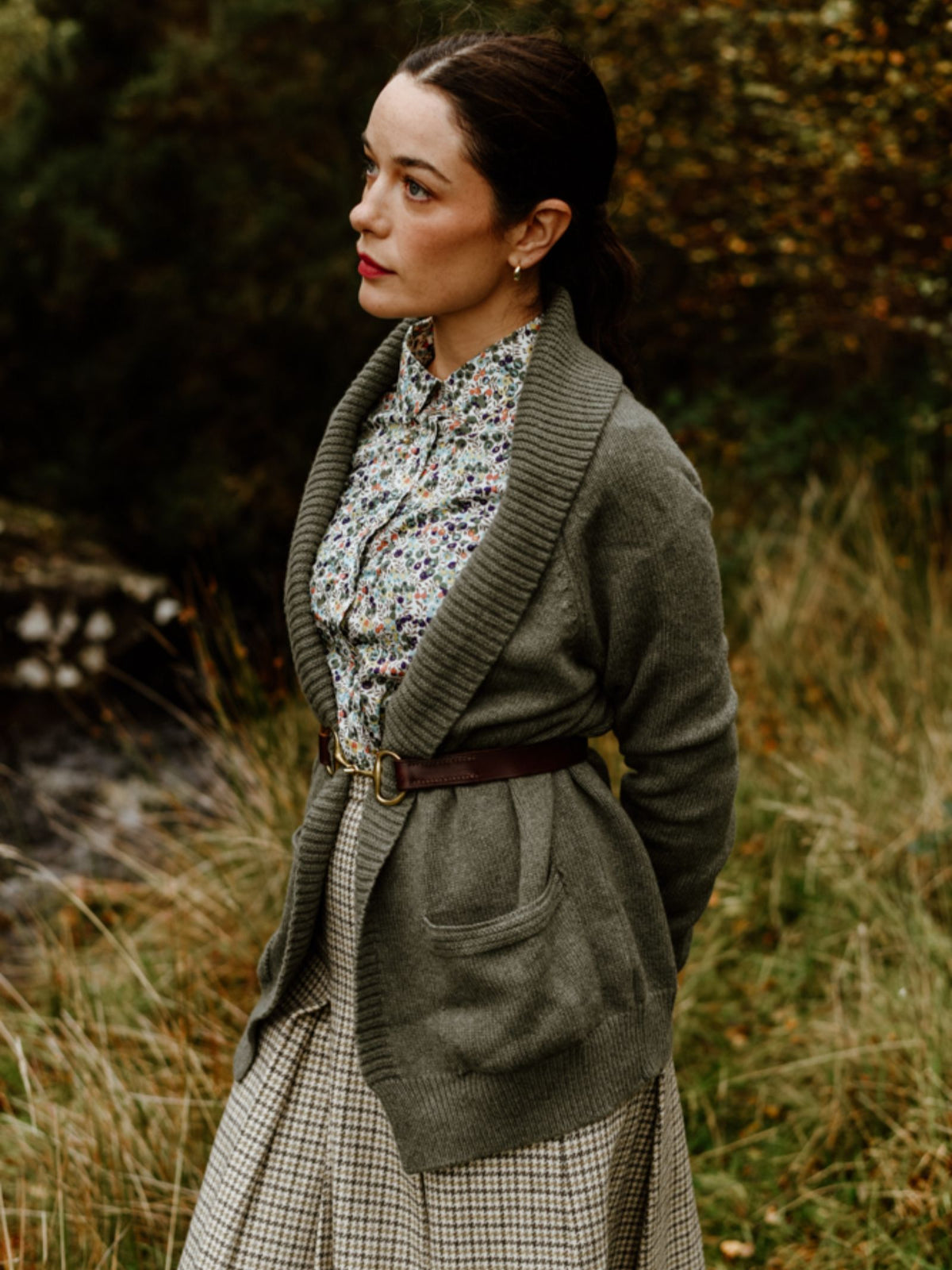 A woman stands outdoors in tall grass wearing a floral blouse, plaid skirt, and a belted Cashmere Shawl Cardigan by Campbell’s of Beauly. Her dark hair is pulled back as she gazes thoughtfully amidst autumn foliage.