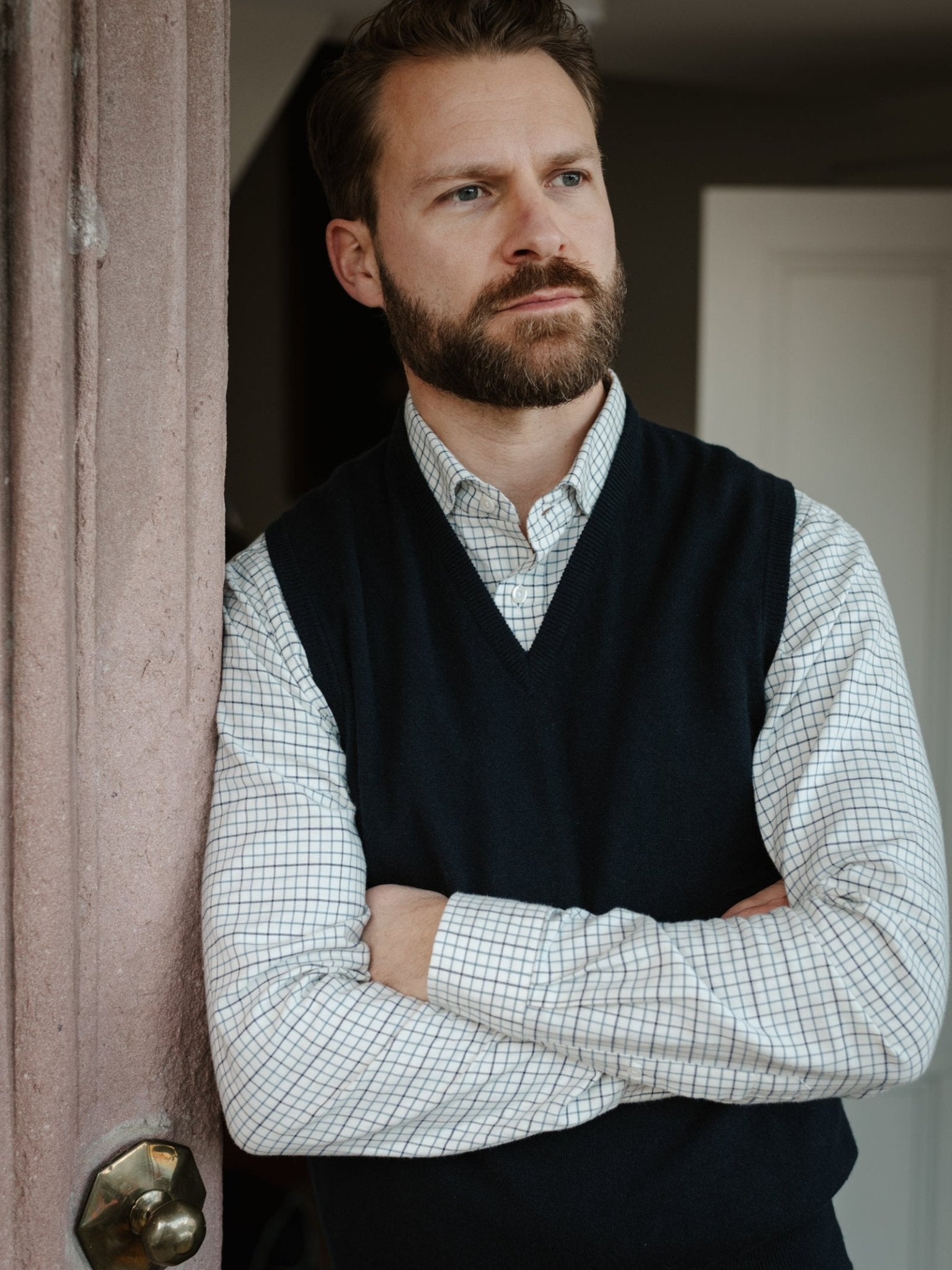 A bearded man stands indoors, leaning against a pink wall with arms crossed, wearing a checked shirt and the classic-fit Cashmere Slipover by Campbell's of Beauly, made in Scotland, as he gazes thoughtfully into the distance.