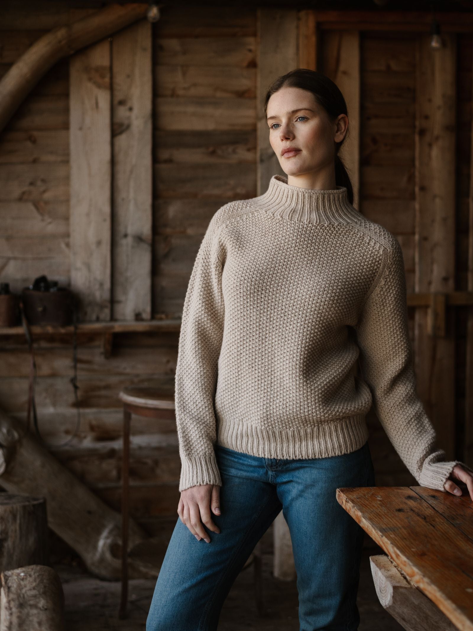A woman in a Campbell's of Beauly Chunky Moss Stitch Polo and blue jeans stands by a wooden table in a rustic cabin with wooden walls, gazing thoughtfully into the distance.