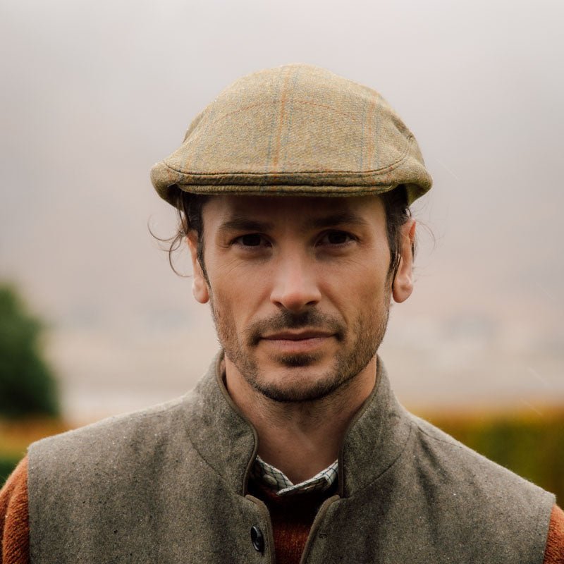 A man with wavy brown hair wearing a flat cap and a buttoned wool vest stands outdoors on a cloudy day, with a soft-focus landscape in the background.
