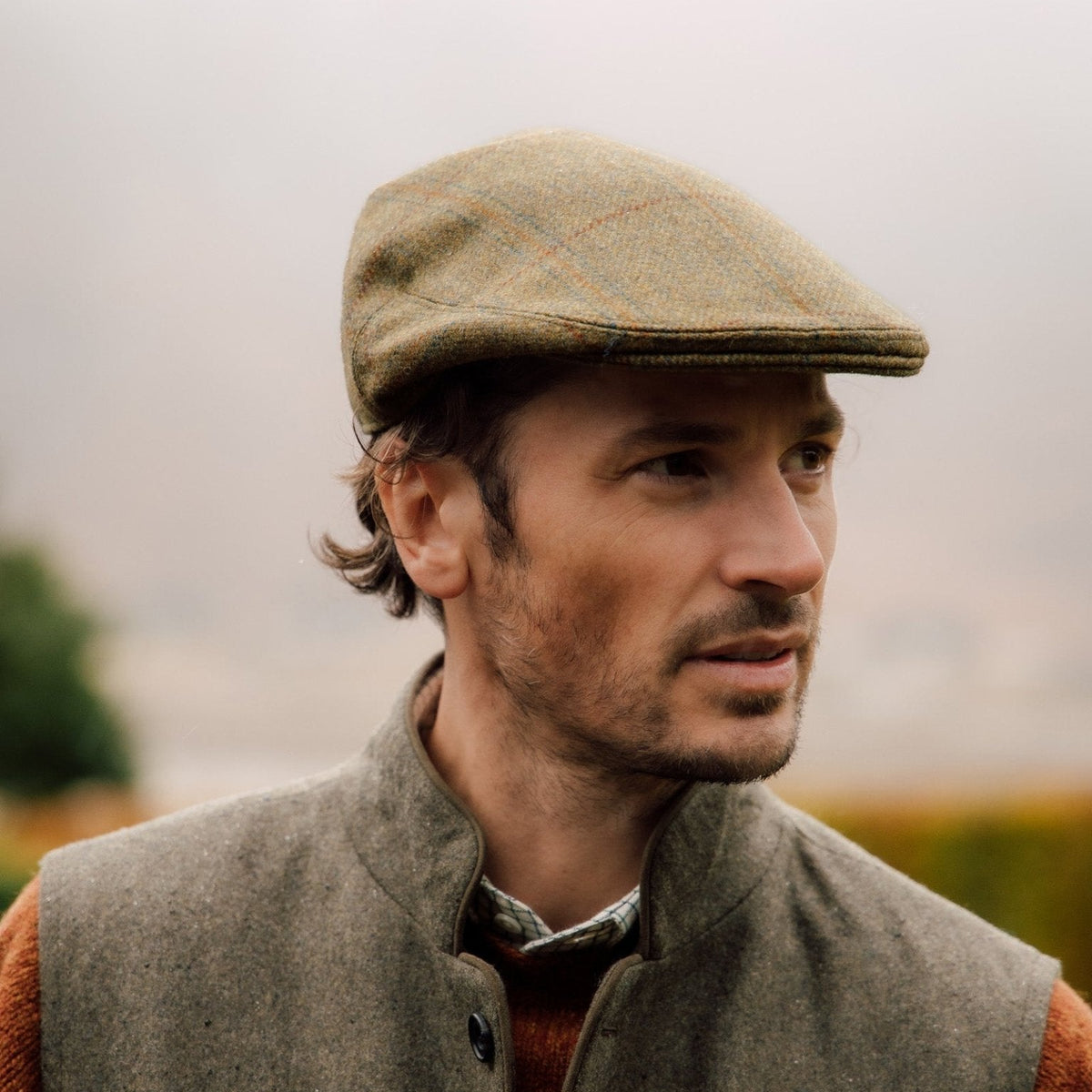 A man in a Green Conon Cap by Campbells of Beauly, paired with a gray vest and rust sweater, stands outdoors with a neutral expression, the blurred background evoking timeless British tweed style.